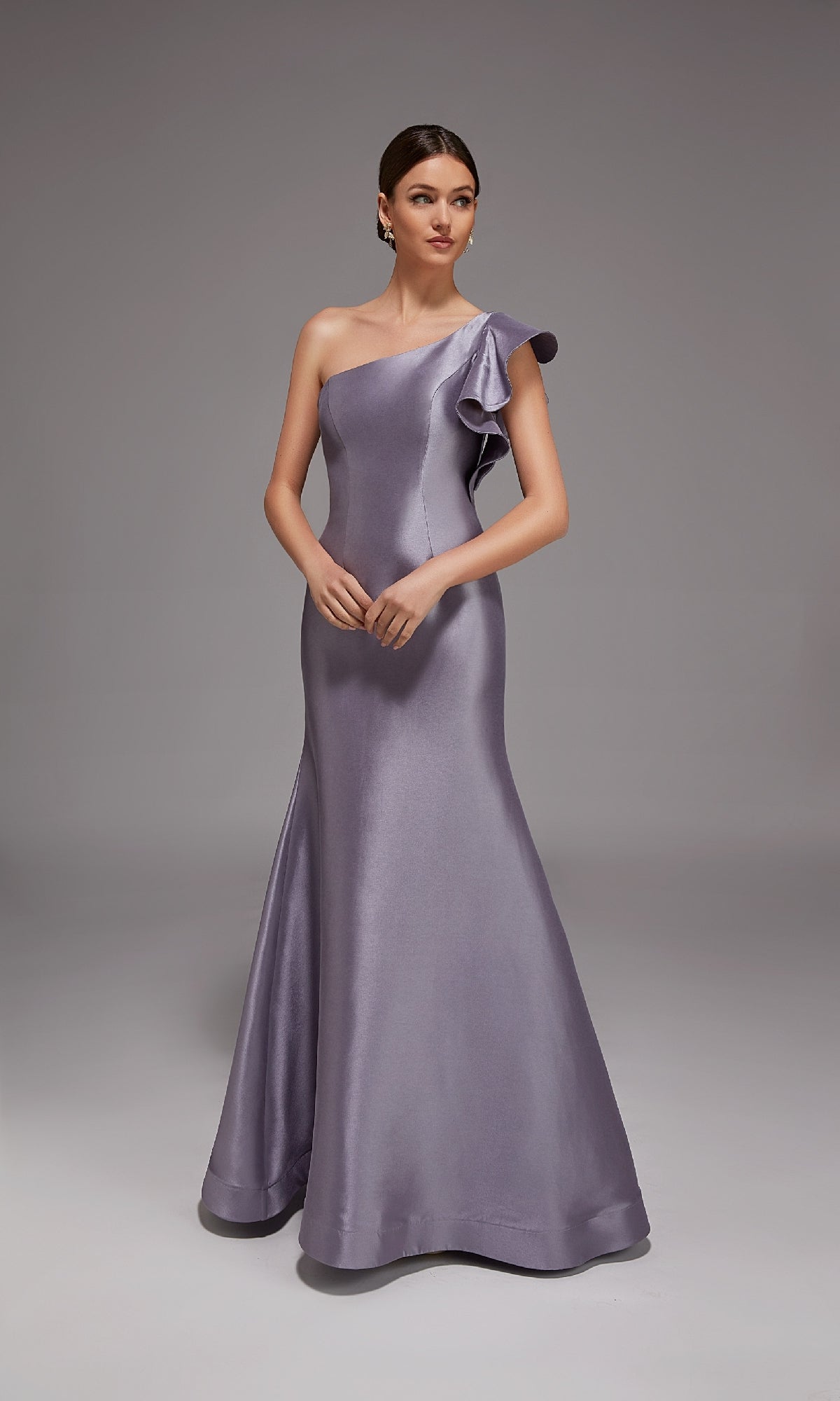 A woman in a floor-length, one-shoulder Lilac Grey Alyce 27826 formal dress with a ruffled shoulder detail stands against a plain gray background.